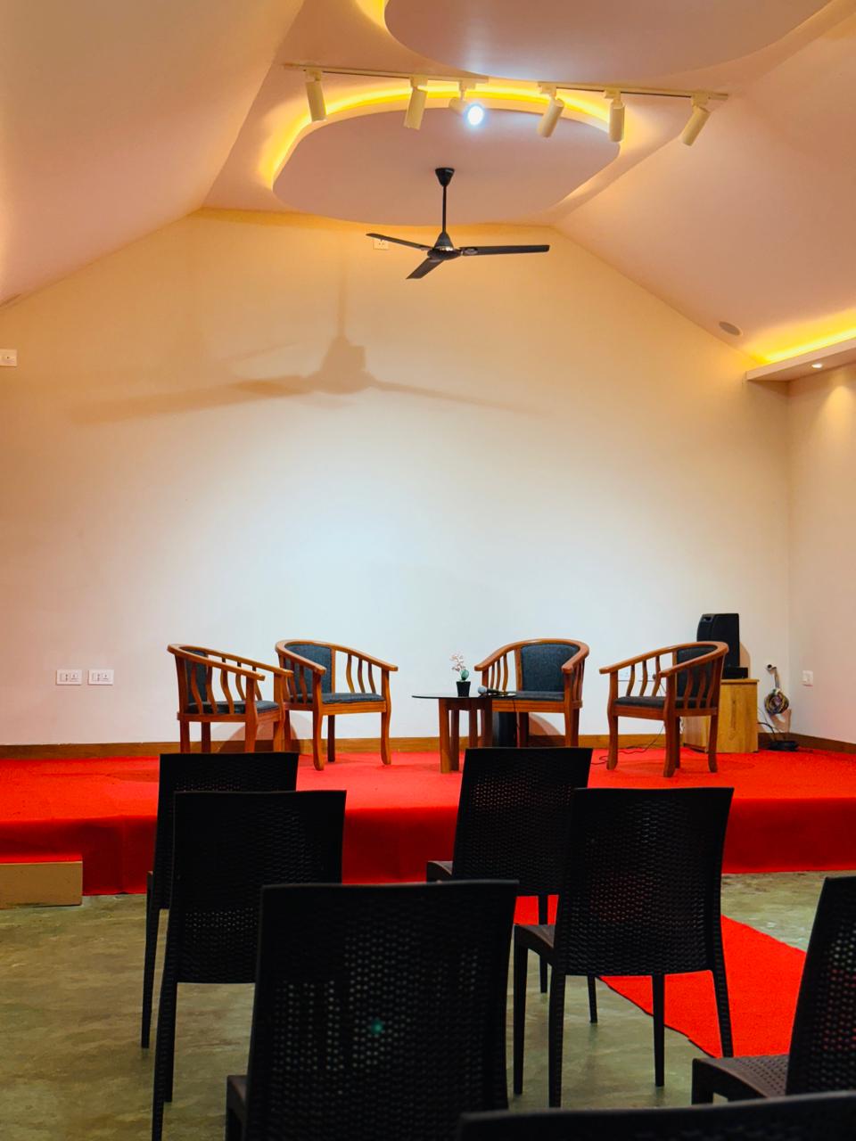 Lounge area with curved blue and orange seating and bean bags facing a large projection screen showing a Crash Bandicoot video game menu. Pink accent lighting streams from tall windows, creating a vibrant, playful environment ideal for socializing or gaming. No visible text. The mood is energetic and inviting.