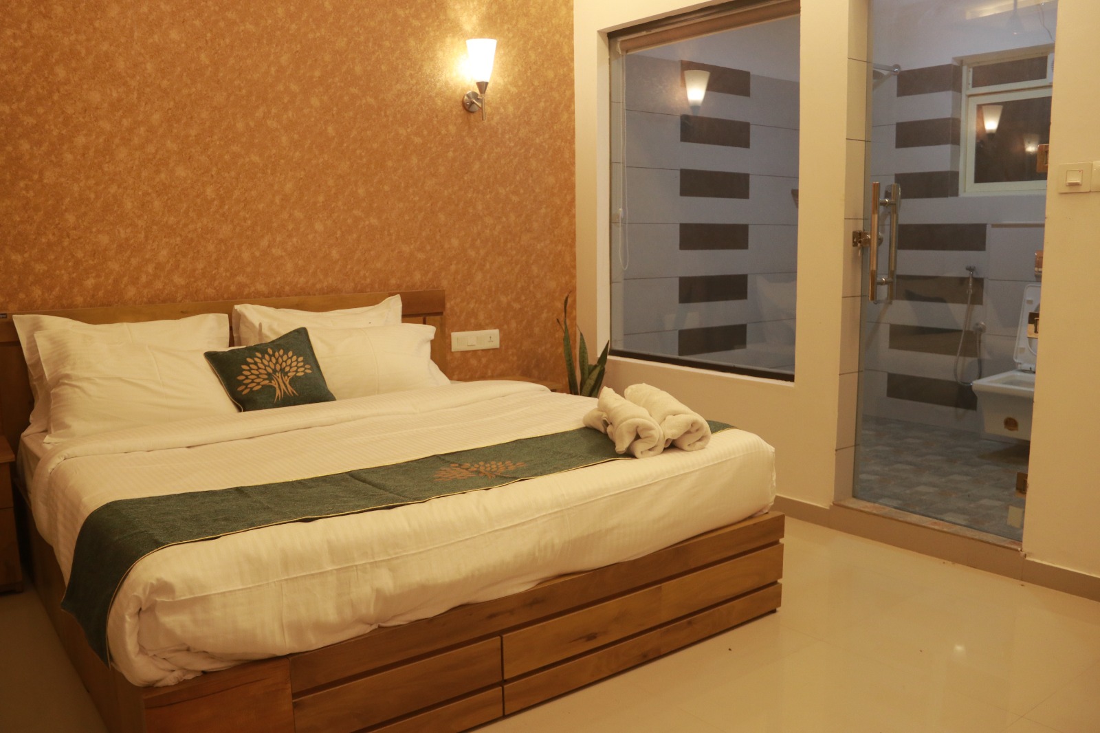 Spacious hotel room featuring a large bed with crisp white linens, green accent runner and pillow, two rolled towels placed on the bed, warm ambient lighting, wooden furniture, and a modern bathroom visible through a glass door. The setting conveys comfort and tranquility. No visible text in the image.