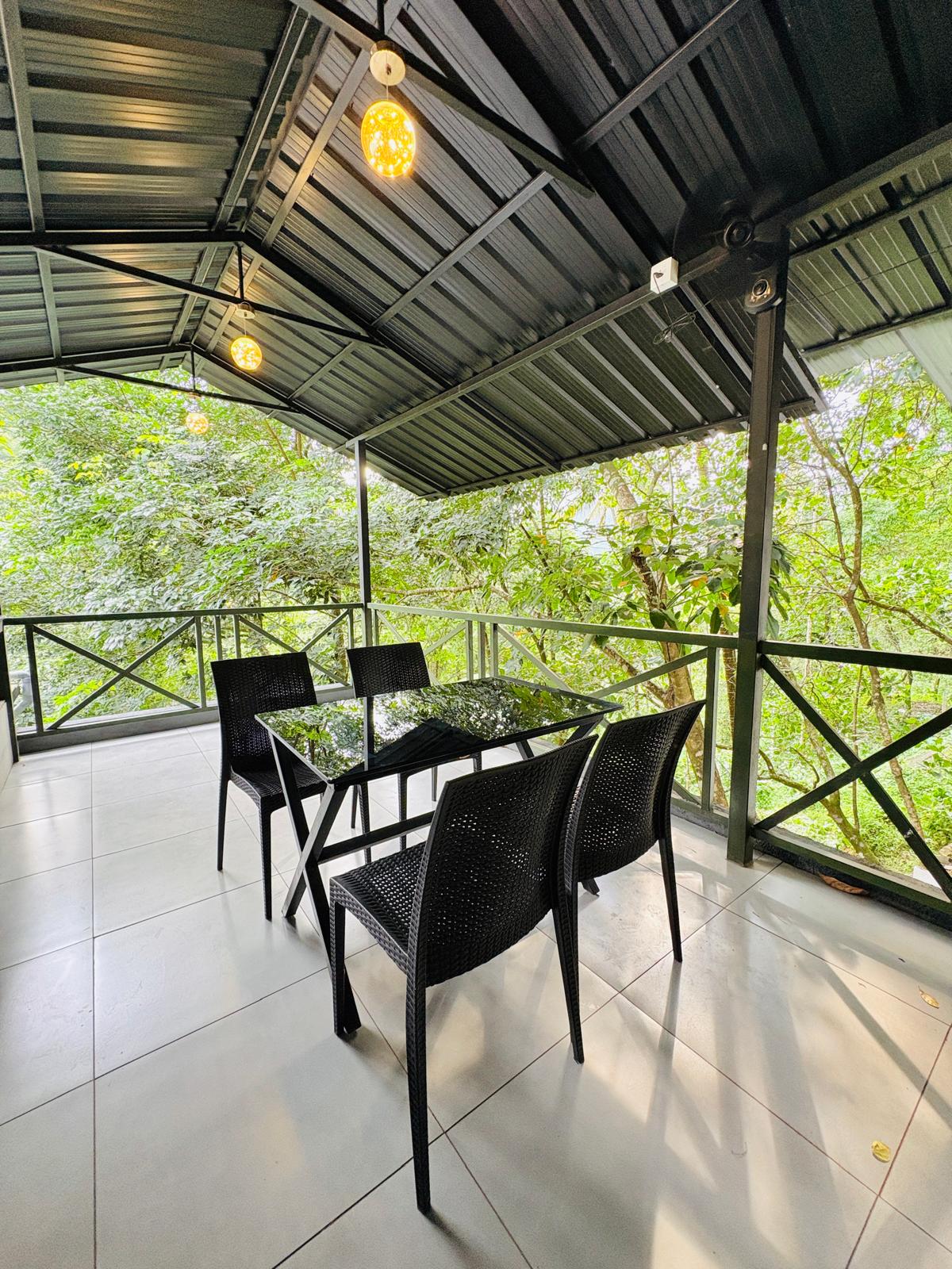 Outdoor dining area at JKR Gardens with wooden tables and chairs arranged on a stone patio, surrounded by lush tropical plants and soft evening lighting. Guests are enjoying meals in a relaxed, cheerful atmosphere with greenery and lanterns enhancing the tranquil setting. No visible text in the image.