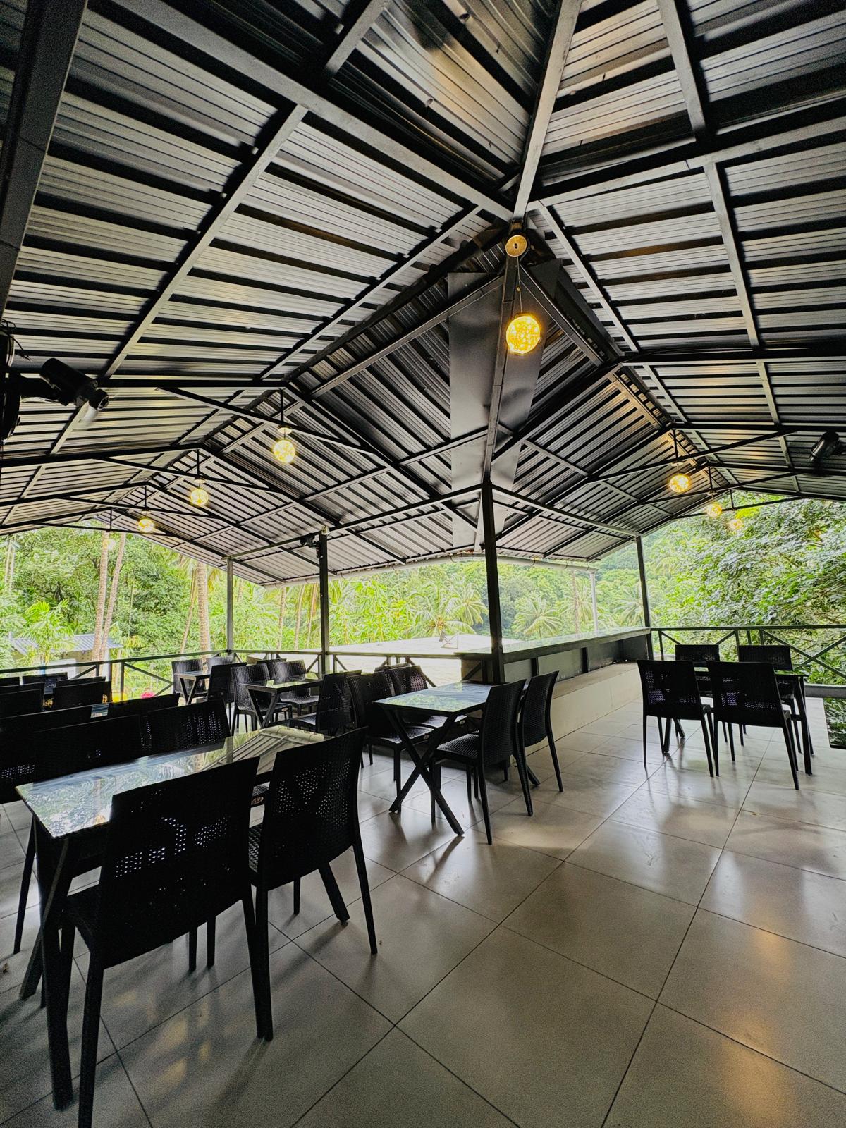 Elegant restaurant interior at JKR Gardens with neatly arranged tables covered in white linens, comfortable wooden chairs, soft ambient lighting, and large windows revealing lush greenery outside. The atmosphere is calm and inviting, ideal for relaxed dining. No visible text in the image.