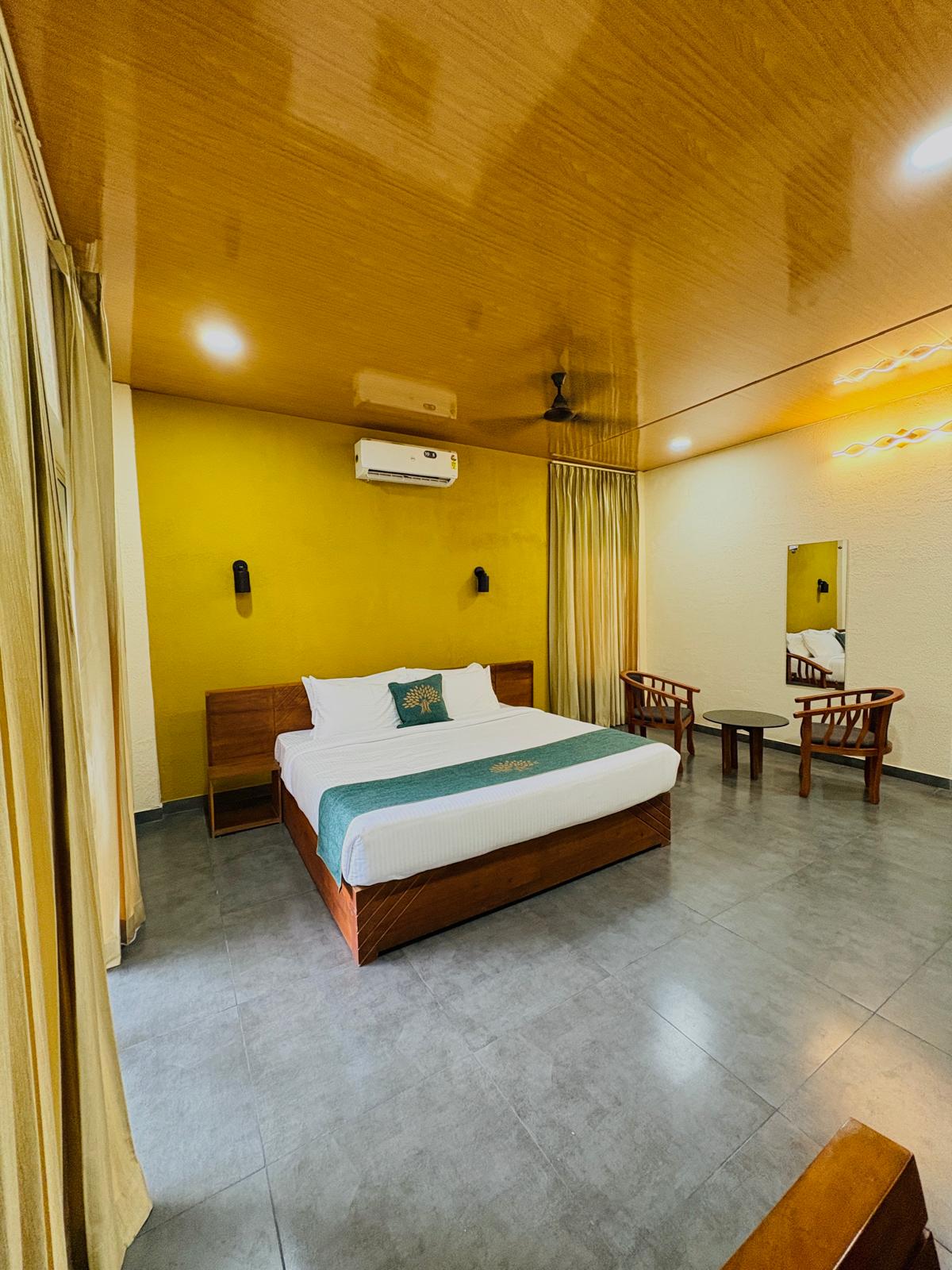 Spacious modern hotel room at JKR Gardens featuring a king-size bed with crisp white linens and a teal runner, wooden furniture, and floor-to-ceiling curtains allowing natural daylight. The room includes a tiled floor, yellow accent wall, air conditioning, and a window overlooking lush green trees, creating a peaceful and inviting atmosphere ideal for relaxation. No visible text.