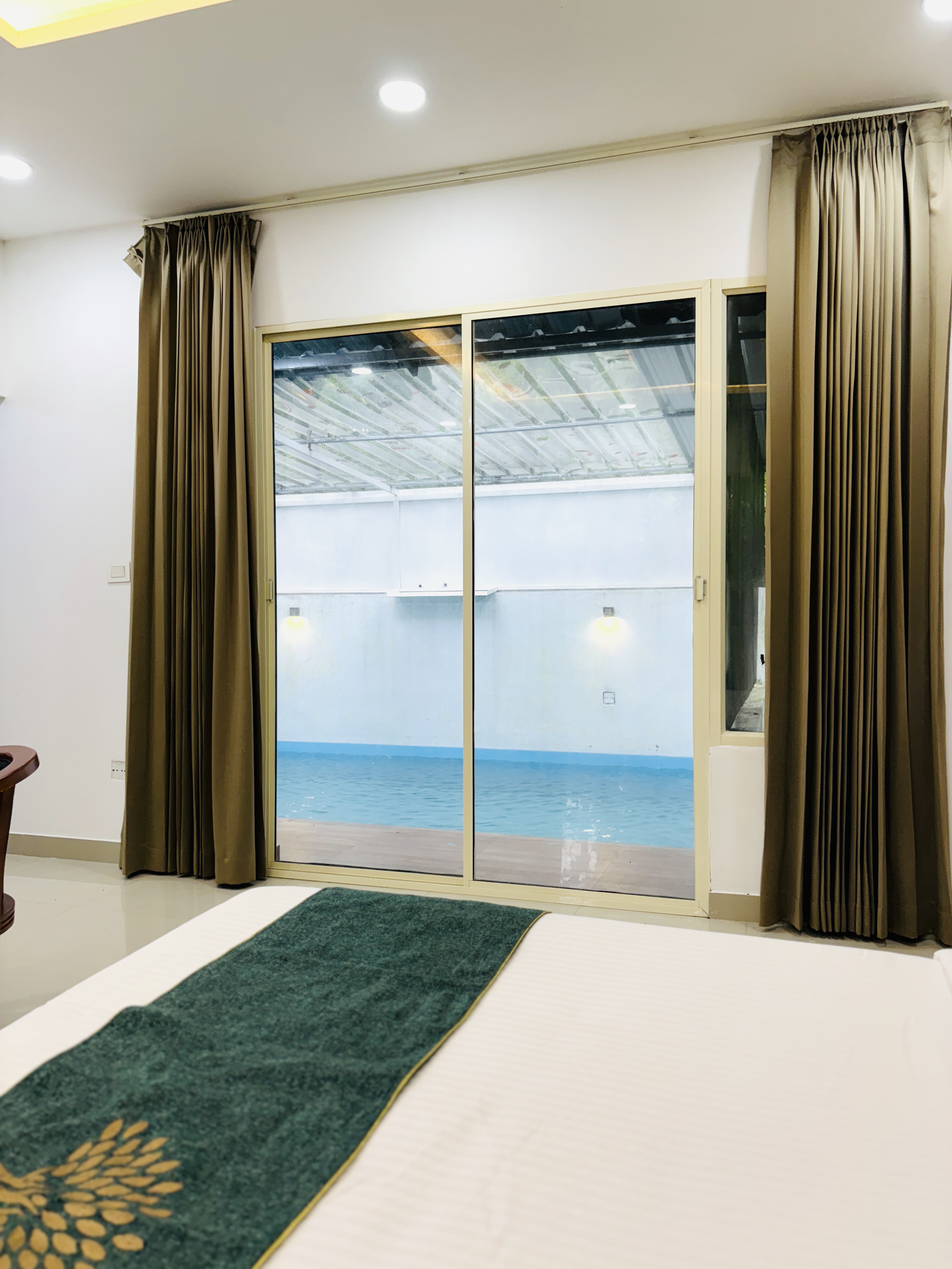 Spacious modern hotel room at JKR Gardens featuring a king-size bed with crisp white linens and a teal runner, wooden furniture, and floor-to-ceiling curtains allowing natural daylight. The room includes a tiled floor, yellow accent wall, air conditioning, and a window overlooking lush green trees, creating a peaceful and inviting atmosphere ideal for relaxation. No visible text.
