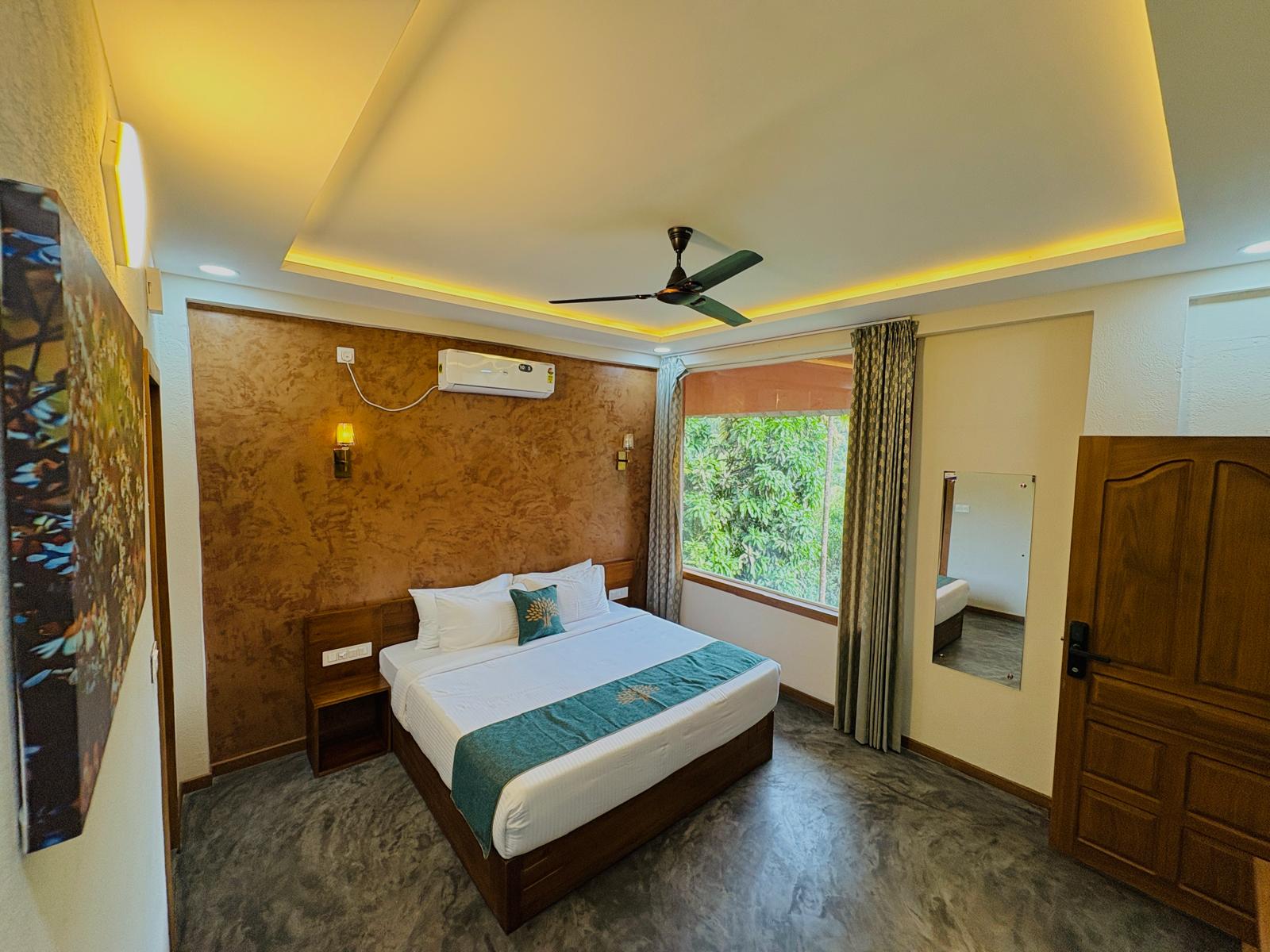 Spacious modern hotel room at JKR Gardens featuring a king-size bed with crisp white linens and a teal runner, wooden furniture, and floor-to-ceiling curtains allowing natural daylight. The room includes a tiled floor, yellow accent wall, air conditioning, and a window overlooking lush green trees, creating a peaceful and inviting atmosphere ideal for relaxation. No visible text.